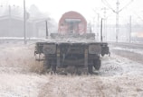 A weathered and abandoned railway flatcar sits in an overgrown rail yard. The flatcar is covered with dry twigs and debris, and the surroundings are misty with a layer of frost on the ground. In the background, there are rail tracks, electrical poles, and a few buildings shrouded in fog.