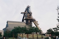 A large industrial structure with a tall chimney and intricate steel framework dominates the scene. It is surrounded by trees and grass, suggesting an industrial site repurposed for public use. People can be seen walking and enjoying the open space, indicating a park-like atmosphere.