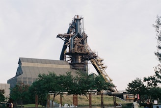 A large industrial structure with a tall chimney and intricate steel framework dominates the scene. It is surrounded by trees and grass, suggesting an industrial site repurposed for public use. People can be seen walking and enjoying the open space, indicating a park-like atmosphere.