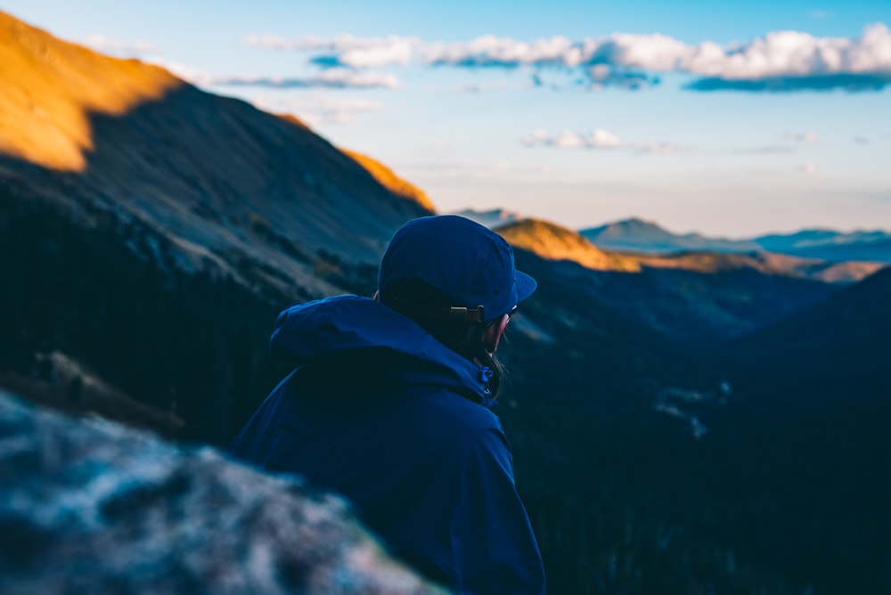 person wearing blue hoodie and blue fitted cap sitting near cliff looking at mountain range under white clouds during daytime