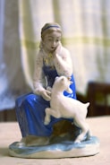A porcelain figurine of a woman in traditional attire, wearing a blue dress and white headscarf, seated with a playful lamb at her side. The figurine is detailed with delicate facial features and a serene expression.