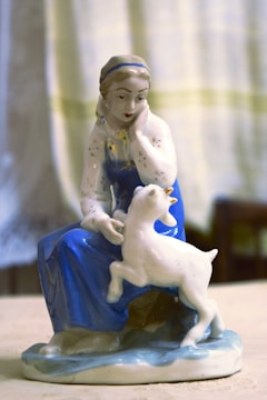 A porcelain figurine of a woman in traditional attire, wearing a blue dress and white headscarf, seated with a playful lamb at her side. The figurine is detailed with delicate facial features and a serene expression.