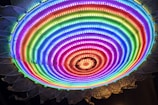 A vibrant circular LED light display with multicolored concentric rings, emitting a bright and colorful glow. The lights transition smoothly from one color to another, creating a rainbow pattern. The outer edge displays a design resembling petals or a sunburst.