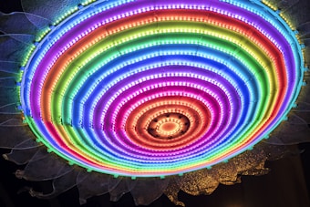 A vibrant circular LED light display with multicolored concentric rings, emitting a bright and colorful glow. The lights transition smoothly from one color to another, creating a rainbow pattern. The outer edge displays a design resembling petals or a sunburst.