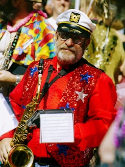 A man wearing a captain's hat and glasses holds a saxophone decorated with colorful sequins, dressed in a bright red sequined vest with star patterns over a red shirt. He stands among people in vibrant attire, suggesting a festive or parade atmosphere.