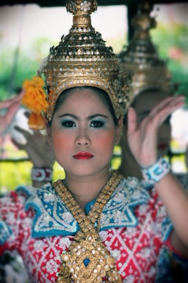 A person is wearing traditional attire with intricate golden headdress adorned with what appears to be pearls. The attire includes a blue and red patterned top with detailed embroidery. The person wears a serious expression and is surrounded by a soft, blurred background.