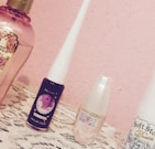 A collection of beauty products is displayed on a lace-covered surface with a pink wall in the background. The items include perfume and cosmetic bottles, each with distinct labels and designs.