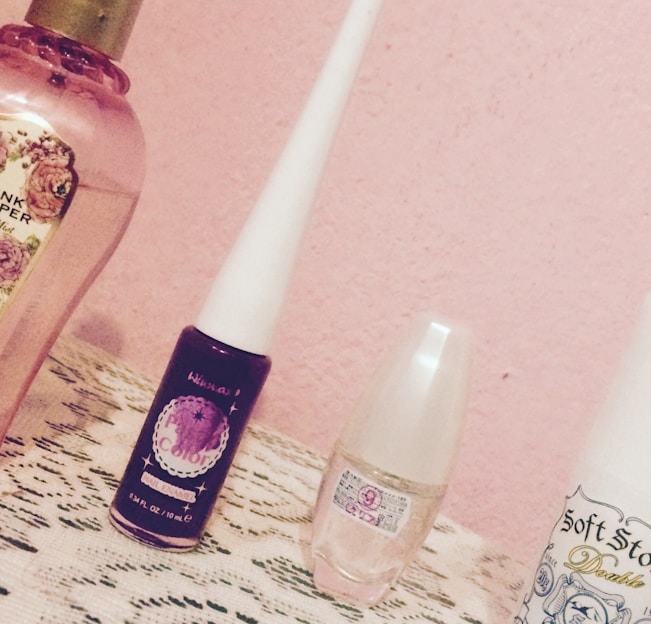 A collection of beauty products is displayed on a lace-covered surface with a pink wall in the background. The items include perfume and cosmetic bottles, each with distinct labels and designs.