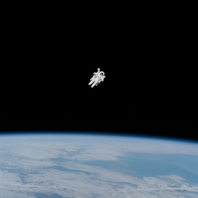 An astronaut is floating in space above the Earth, surrounded by a vast expanse of darkness. The Earth's surface is visible below, showing a thin blue atmosphere and cloud formations.