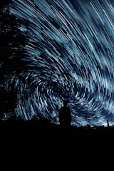 A silhouette of a person stands against a backdrop of swirling, streaked stars in the night sky. The long exposure creates a sense of movement and vortex-like patterns among the stars. Trees frame the scene at the edges, adding depth and contrast to the composition.