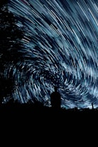 A silhouette of a person stands against a backdrop of swirling, streaked stars in the night sky. The long exposure creates a sense of movement and vortex-like patterns among the stars. Trees frame the scene at the edges, adding depth and contrast to the composition.