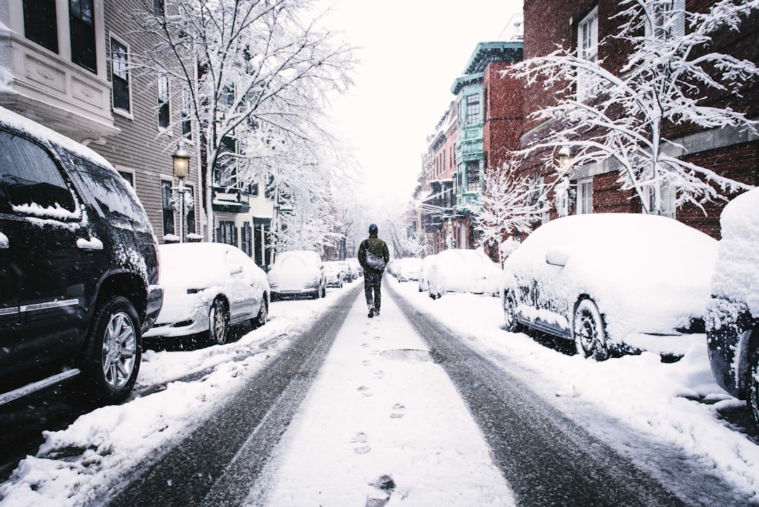 man walking on road covered with snow between vehicles and concrete buildings during daytime