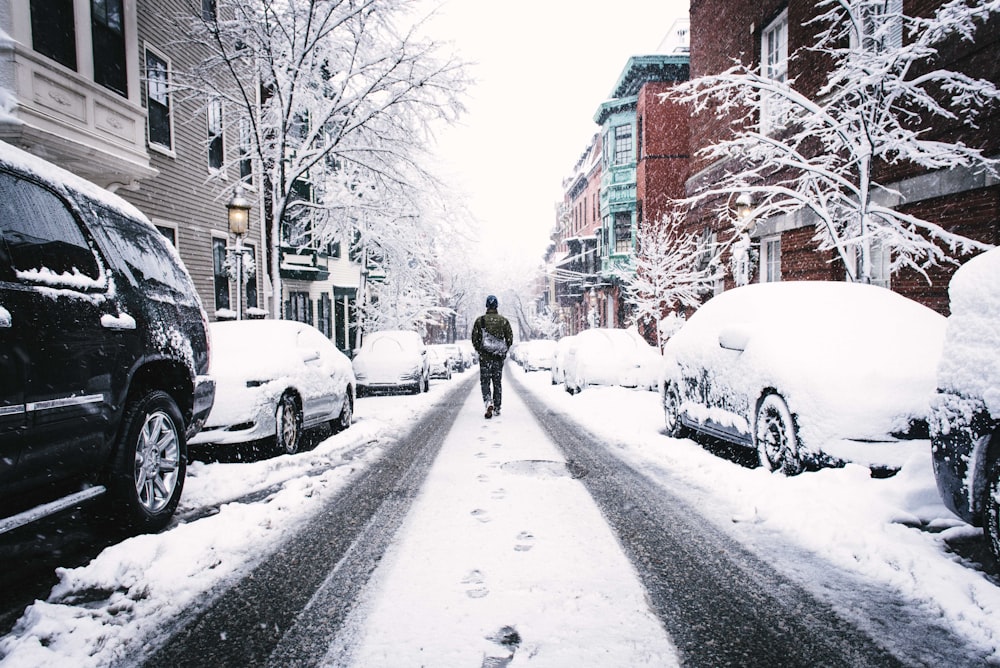 uomo che cammina sulla strada coperta di neve tra i veicoli e gli edifici in cemento durante il giorno