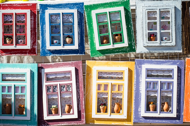 Colorful and artistic window frames arranged in a grid, each frame features a unique design with different arrangements of lace curtains, miniature pots, and decorative items.