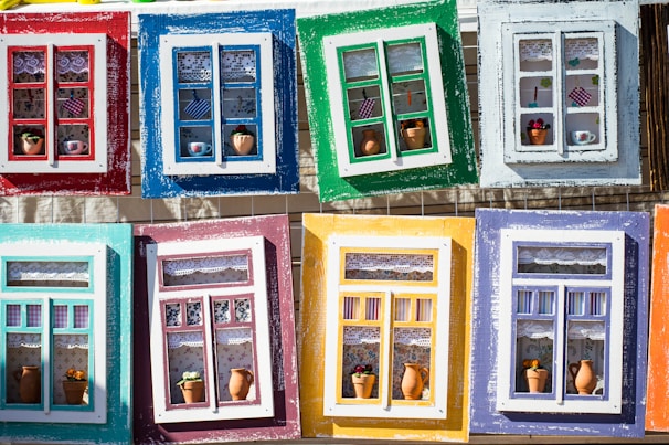Colorful and artistic window frames arranged in a grid, each frame features a unique design with different arrangements of lace curtains, miniature pots, and decorative items.