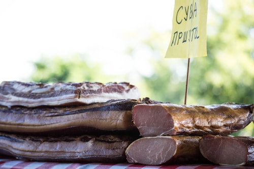Several stacked slices of cured meat displayed on a table with a yellow sign featuring Cyrillic writing. The background is a blurred green landscape, likely trees, indicating an outdoor setting.
