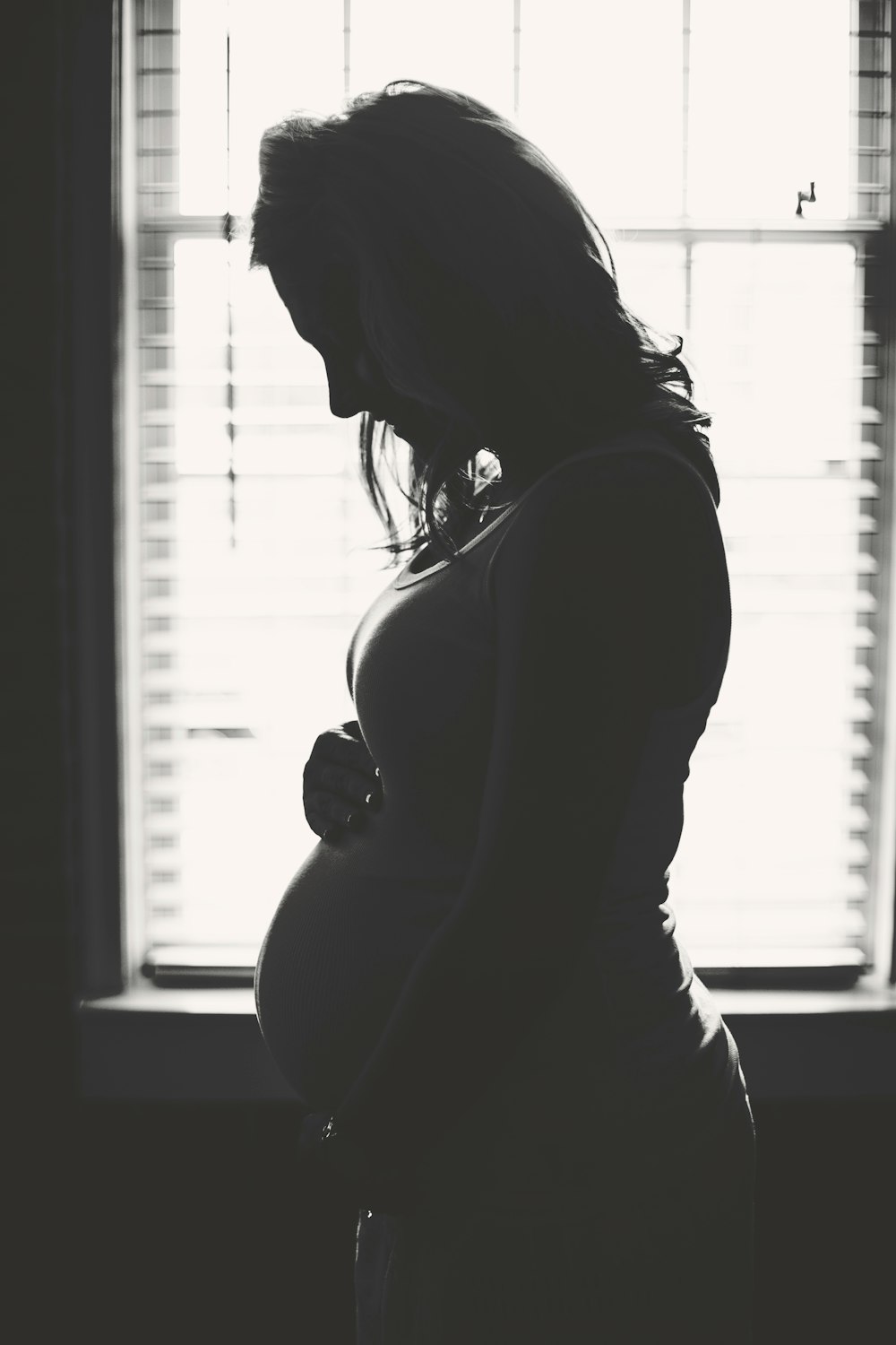 A black and white silhouette of a pregnant woman looking down and holding her belly in front of a window.