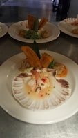 A gourmet dish presented on a large white plate, featuring a scallop shell at the forefront filled with a seafood ceviche, garnished with herbs and small diced vegetables. Behind the shell, there are two plantain chips standing upright. The background shows additional plates with similar gourmet arrangements.