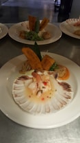 A gourmet dish presented on a large white plate, featuring a scallop shell at the forefront filled with a seafood ceviche, garnished with herbs and small diced vegetables. Behind the shell, there are two plantain chips standing upright. The background shows additional plates with similar gourmet arrangements.