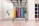 A neatly organized rack holds colorful hexagonal weights arranged in rows with varying colors including yellow, orange, purple, blue, and green. The setting appears to be a clean, well-lit gym environment with other fitness equipment like resistance bands hanging on the mirror-lined wall. A set of shelves on the right contains rolled-up yoga mats.