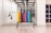 A neatly organized rack holds colorful hexagonal weights arranged in rows with varying colors including yellow, orange, purple, blue, and green. The setting appears to be a clean, well-lit gym environment with other fitness equipment like resistance bands hanging on the mirror-lined wall. A set of shelves on the right contains rolled-up yoga mats.