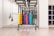 A neatly organized rack holds colorful hexagonal weights arranged in rows with varying colors including yellow, orange, purple, blue, and green. The setting appears to be a clean, well-lit gym environment with other fitness equipment like resistance bands hanging on the mirror-lined wall. A set of shelves on the right contains rolled-up yoga mats.