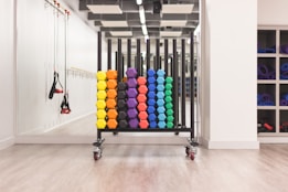 A neatly organized rack holds colorful hexagonal weights arranged in rows with varying colors including yellow, orange, purple, blue, and green. The setting appears to be a clean, well-lit gym environment with other fitness equipment like resistance bands hanging on the mirror-lined wall. A set of shelves on the right contains rolled-up yoga mats.