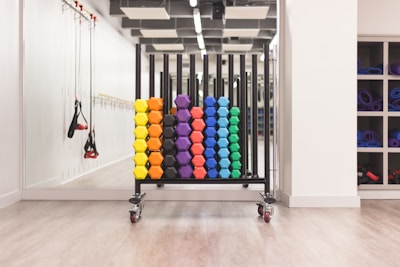 A neatly organized rack holds colorful hexagonal weights arranged in rows with varying colors including yellow, orange, purple, blue, and green. The setting appears to be a clean, well-lit gym environment with other fitness equipment like resistance bands hanging on the mirror-lined wall. A set of shelves on the right contains rolled-up yoga mats.