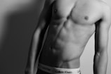 A black and white photograph of a male torso focusing on the abdominal muscles. The person is leaning slightly to one side, showcasing defined muscle tone. The waistband of Calvin Klein underwear is visible, adding a sense of brand identity to the image.