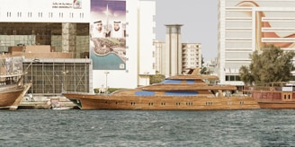 A luxurious wooden yacht is docked in a vibrant city waterfront area. The scene includes buildings in the background, featuring advertisements and signs in Arabic. The yacht has sleek, elegant lines with several windows, and there are other smaller boats present. The water is calm, reflecting the structures.