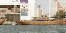 A luxurious wooden yacht is docked in a vibrant city waterfront area. The scene includes buildings in the background, featuring advertisements and signs in Arabic. The yacht has sleek, elegant lines with several windows, and there are other smaller boats present. The water is calm, reflecting the structures.