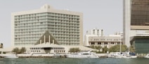 A large, modern hotel building with a distinctive triangular entrance, located along a waterfront with several yachts docked in the foreground. The architecture features tall glass windows, and the surrounding area includes palm trees and a few other buildings.