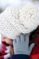 A close-up view of a person wearing a thick, textured white knit beanie with a pom-pom. The individual is also wearing gray gloves and a red scarf, suggesting a cold, wintery environment.