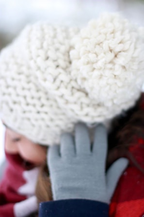 A close-up view of a person wearing a thick, textured white knit beanie with a pom-pom. The individual is also wearing gray gloves and a red scarf, suggesting a cold, wintery environment.