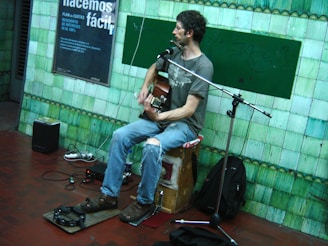 A person is sitting on a wooden box playing a guitar, equipped with a microphone and amplifiers, set against a green tiled wall in an indoor space. The person is wearing a casual outfit with jeans and a T-shirt.