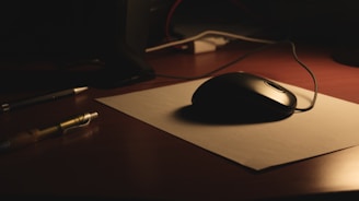 A dimly lit desk with a black computer mouse resting on a piece of white paper. Nearby, a pen lies on the wooden surface. Cables are slightly visible in the background, suggesting an electronic setup.
