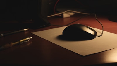 A dimly lit desk with a black computer mouse resting on a piece of white paper. Nearby, a pen lies on the wooden surface. Cables are slightly visible in the background, suggesting an electronic setup.