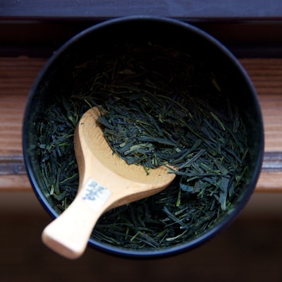 A wooden spoon rests in a black bowl filled with vibrant green tea leaves. The texture of the leaves is visible, and the spoon has characters written on its handle.