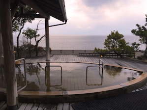A serene onsen setting with a circular hot spring pool surrounded by a wooden deck overlooking the ocean. The pool is bordered by handrails, and there are trees providing a natural atmosphere. The overcast sky adds a calming effect to the scene.
