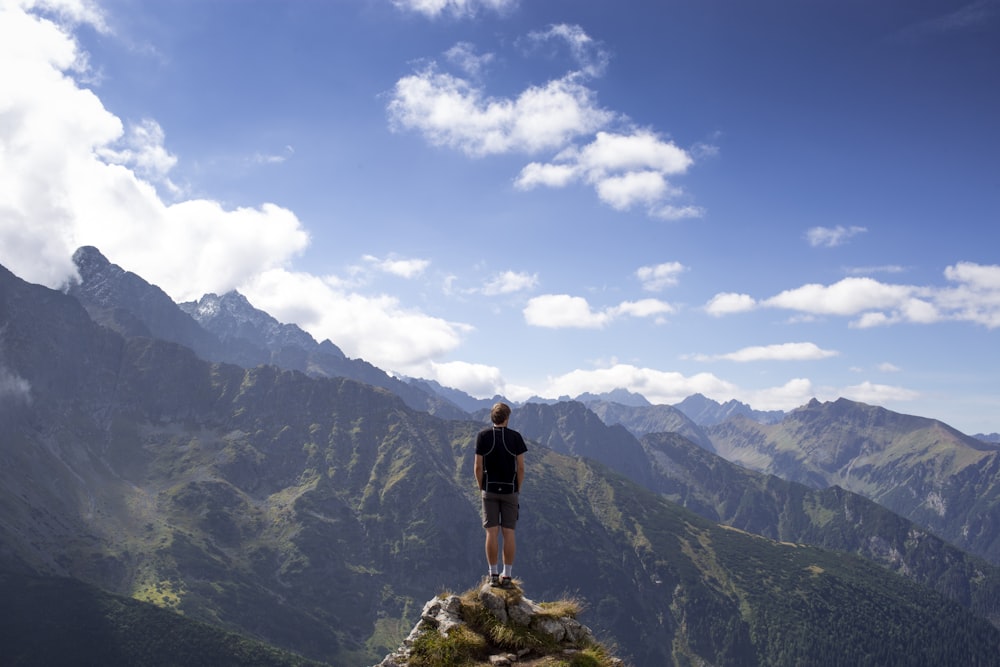 hombre con camisa negra y pantalones cortos grises en la colina de la montaña junto a las montañas bajo cielos nublados blancos y azules