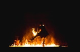 A performer dances amidst flames, creating a dramatic and powerful scene. The fire illuminates the dancer and the surrounding area, casting bright orange and yellow hues against a dark backdrop. The figure appears dynamic and full of motion, enhancing the intensity of the spectacle.