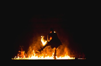 A performer dances amidst flames, creating a dramatic and powerful scene. The fire illuminates the dancer and the surrounding area, casting bright orange and yellow hues against a dark backdrop. The figure appears dynamic and full of motion, enhancing the intensity of the spectacle.