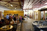 A lively indoor space with groups of people gathered in a cafe-like setting. There are wooden tables and chairs with people engaged in conversation. The environment is open with exposed beams, visible light fixtures, and a mix of rustic and modern elements. Decorative items and potted plants add a cozy atmosphere.