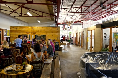 A lively indoor space with groups of people gathered in a cafe-like setting. There are wooden tables and chairs with people engaged in conversation. The environment is open with exposed beams, visible light fixtures, and a mix of rustic and modern elements. Decorative items and potted plants add a cozy atmosphere.