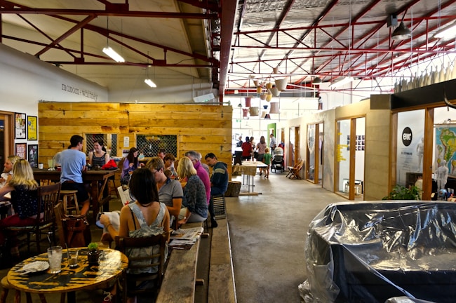 A lively indoor space with groups of people gathered in a cafe-like setting. There are wooden tables and chairs with people engaged in conversation. The environment is open with exposed beams, visible light fixtures, and a mix of rustic and modern elements. Decorative items and potted plants add a cozy atmosphere.