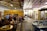 A lively indoor space with groups of people gathered in a cafe-like setting. There are wooden tables and chairs with people engaged in conversation. The environment is open with exposed beams, visible light fixtures, and a mix of rustic and modern elements. Decorative items and potted plants add a cozy atmosphere.