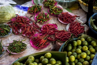 A market stall displays an assortment of fresh produce. Plates filled with vibrant red and green chilies are arranged on a stone surface, alongside baskets of green limes. There is also a large cabbage visible in the background, partially obscured.