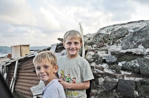 Two young boys are standing in front of a rustic stone wall, both smiling at the camera. The older boy on the right is wearing a T-shirt with a cartoon graphic, and the younger boy on the left is wearing a light-colored shirt. Behind them, there are makeshift structures made from corrugated metal and wood. The sky is overcast with clouds, adding a soft light to the scene.
