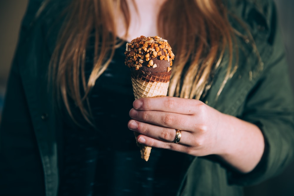 woman in green jacket holding chocolate ice cream coated of nuts during daytime shallow focus photography
