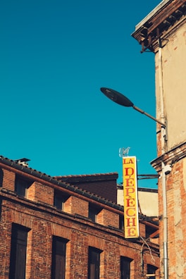 A historic brick building with a pitched roof and several windows is visible. A bright yellow vertical sign with red letters spelling 'LA DÉPÊCHE' is attached to the building. The sky is clear and deep blue, adding contrast and highlighting the architecture.